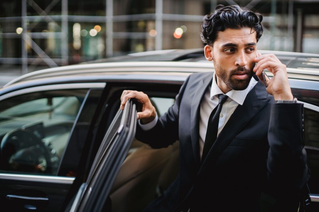 Executive in a dark suit on a phone call steps out of a black car on a city street, a high-risk moment for executive protection in California.