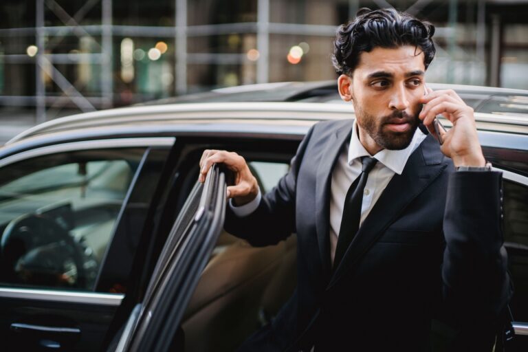 Executive in a dark suit on a phone call steps out of a black car on a city street, a high-risk moment for executive protection in California.